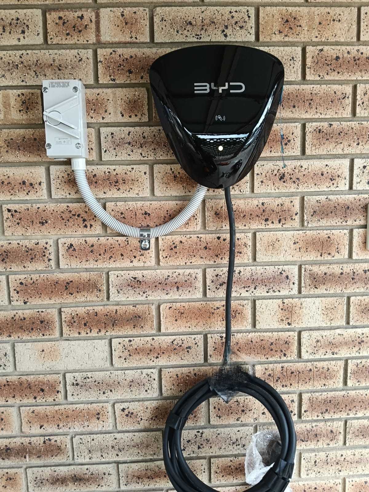 Wall-mounted BYD electric vehicle charger on a brick wall, with a white weatherproof box and a coiled cable beneath.